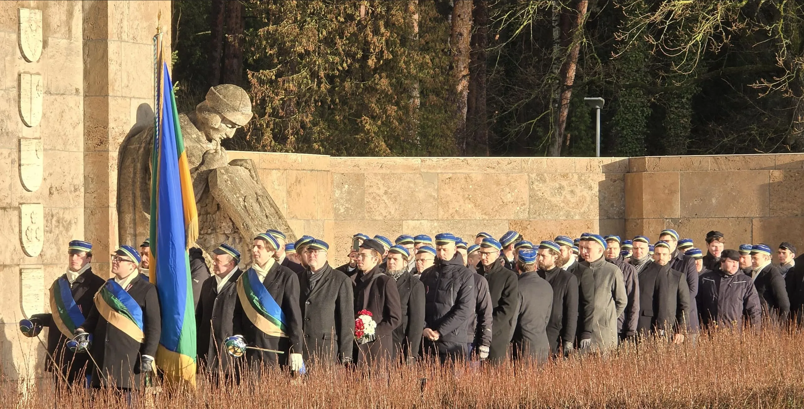 Fraternitas Lettica biedri zilos deķeļos gara ierindā ar korporācijas karogu pie akmens pieminekļa